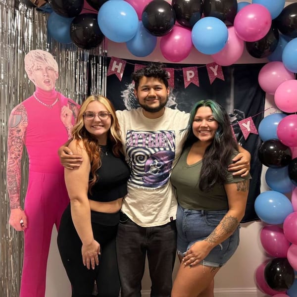 Three smiling people posing with a Machine Gun Kelly cutout and pink, blue, and black balloons.