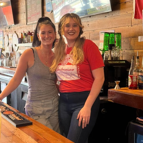 Two smiling women, Commerce Sports Bar and Grill bartenders, stand behind a wooden bar with bottles and glasses.