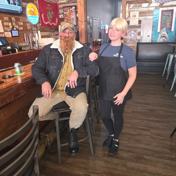 A man with a fake beard sits at the bar, and a woman in an apron stands beside him.